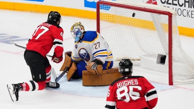 A men's hockey player scores a goal against the opposing goalie as another player looks on.