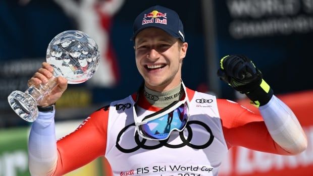 A men's skier - wearing a red and white ski suit, and navy blue Red Bull hat - grasps a Crystal Globe in one hand and pumps the first of her other hand, while posing for a photograph.