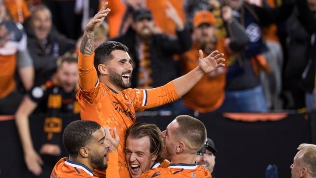 A male soccer player wearing orange uniform is lifted by three teammates as they all celebrate a goal while members of the audience watch on from the back.