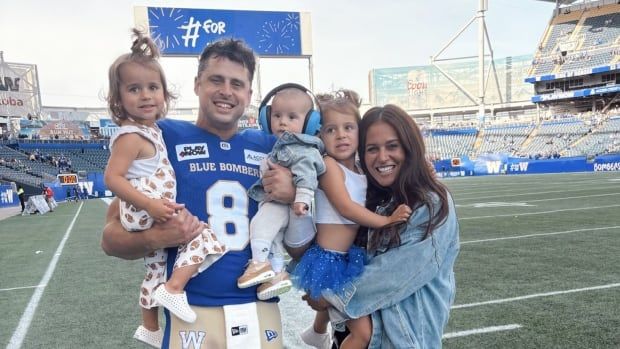 A football player in blue and gold stands on the sidelines holding two young children. Next to him is a woman holding another child.