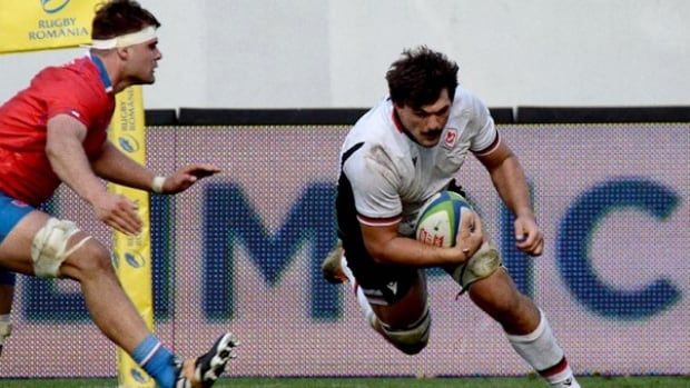 A male rugby player carries the ball past an opposition player.