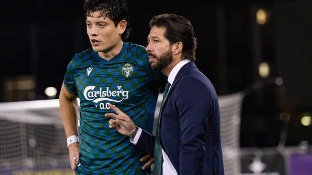 A male soccer coach wearing a suit and tie stands alongside one of his players on the sideline during a match.