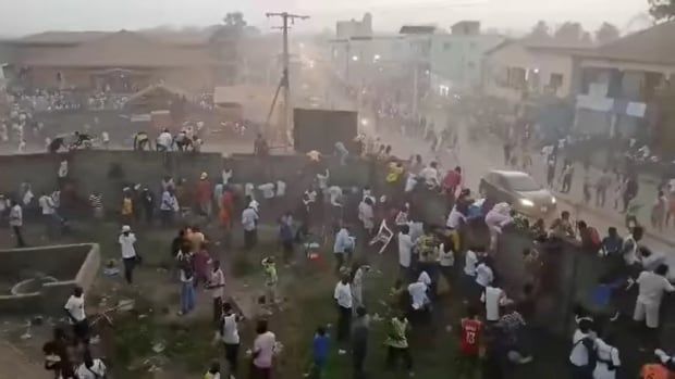 Dozens of people are shown from an elevated view, milling about various parts of a settlement with low-rise buildings and dirt road.