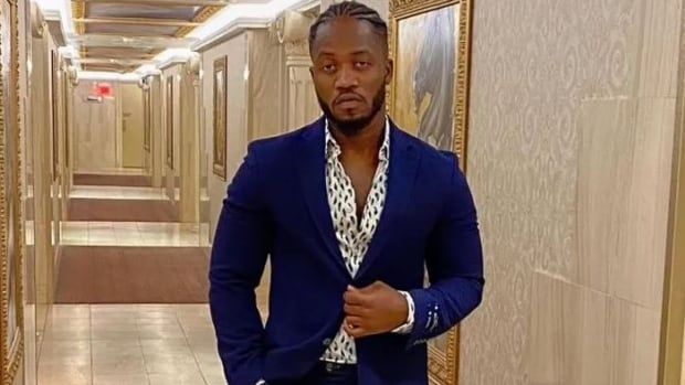 A man in a blue suit stands in a hallway with beige tile walls. 