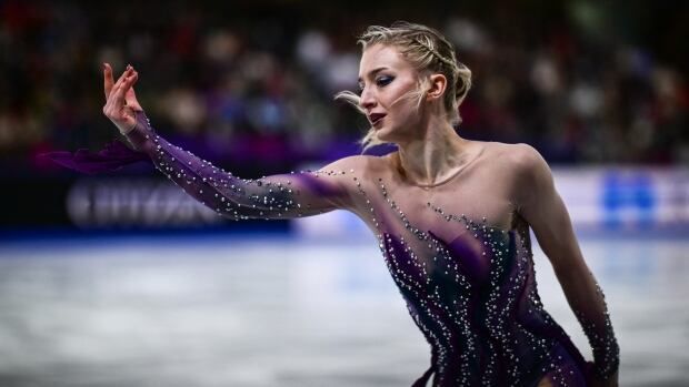 A women's figure skater, wearing a purple outfit with sequins, extends her hand while perofrming.