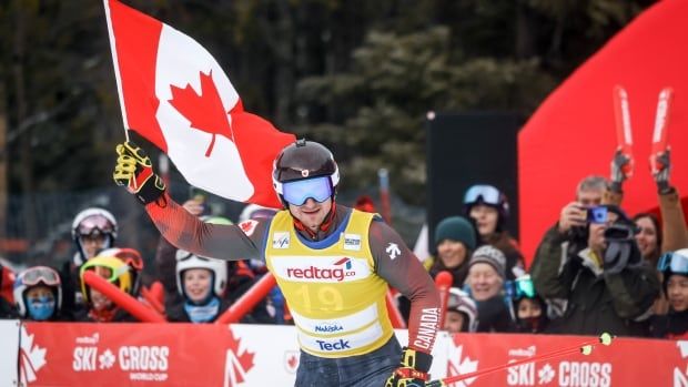 A male skier is seen wearing race gear and holding the Canadian flag as he smiles. Members of the audience watch on behind him. 