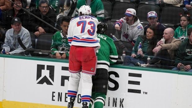 A male ice hockey player leaps into the air as he checks an opposing player into the boards from behind while fans watch from behind the glass.