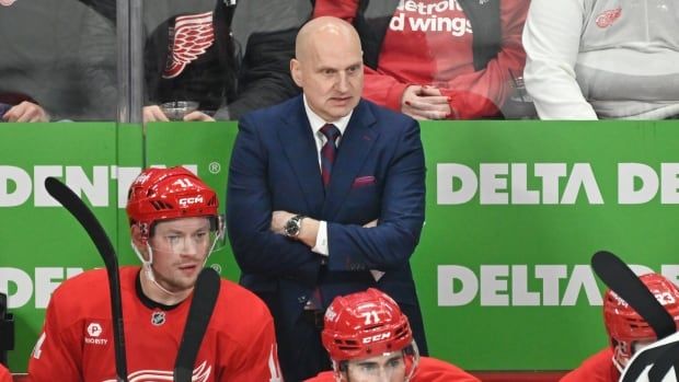 A hockey coach stands with his arms crossed on the bench.