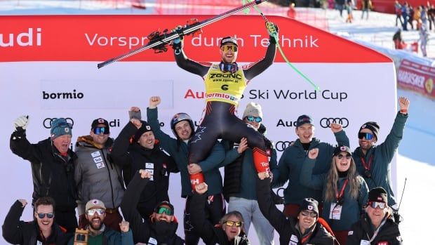 A male alpine skier raises his skis in celebration while being hoisted in the air by his team.