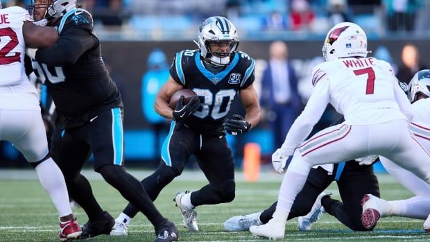 A male football running back wearing number 30 carries the ball in his right hand as a defender wearing number seven closes in.