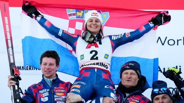 A female alpine skier smiles while raising the Croatian flag behind her back as teammates hoist her into the air.