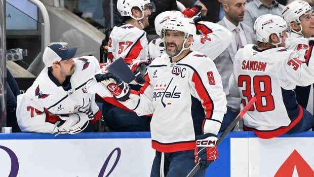 A male ice hockey forward smiles while touching gloves with teammates in celebration as he skates past the bench during a game.