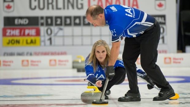 A female curling skip delivers a stone with her right hand as her male mixed doubles partner sweeps ahead of her.