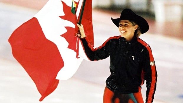 A female speed skater wearing a cowboy hat skates around while waving a Canadian flag with her right hand.