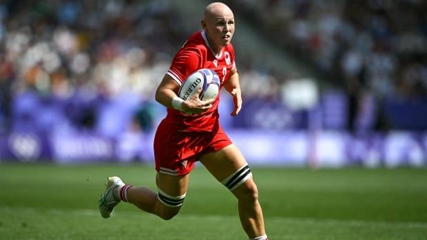 A female rugby sevens player representing Canada runs with the ball in her right hand during a game as fans watch from the stands.