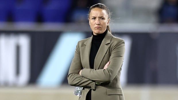 A female soccer coach walks the sideline during a game with her arms crossed.