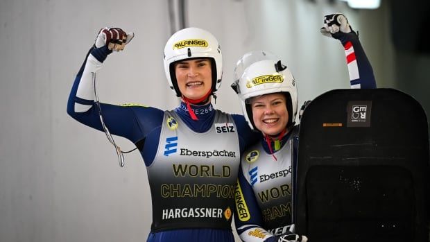 Two women's luge athletes pose for a photograph while smiling and each flexing their biceps on the luge track.