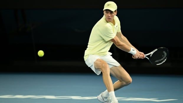 A male tennis player plays a backhand on a hard court.