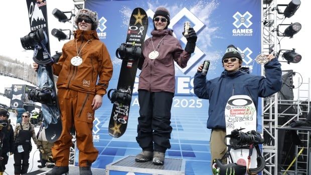 Mark McMorris is standing on a podium outside in a brown snowsuit next to other competitors