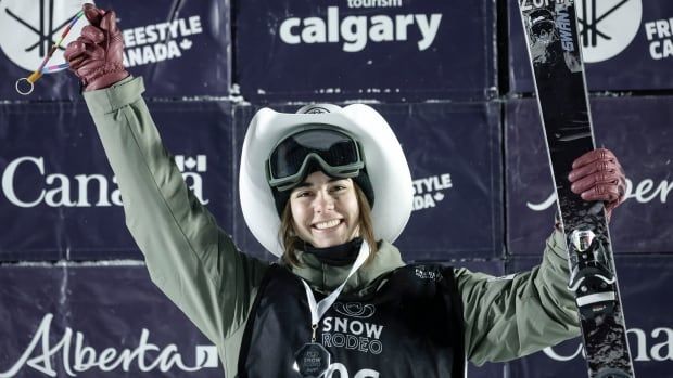 A female freestyle skier wearing a cowboy hat and bronze medal around her neck smiles while raising her arms in celebration as she holds her skies in her left hand on the podium.