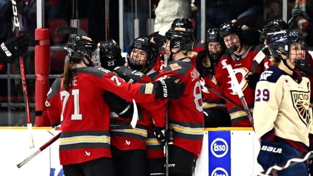 Female ice hockey players wrap their arms around a smiling teammate in celebration during a game.