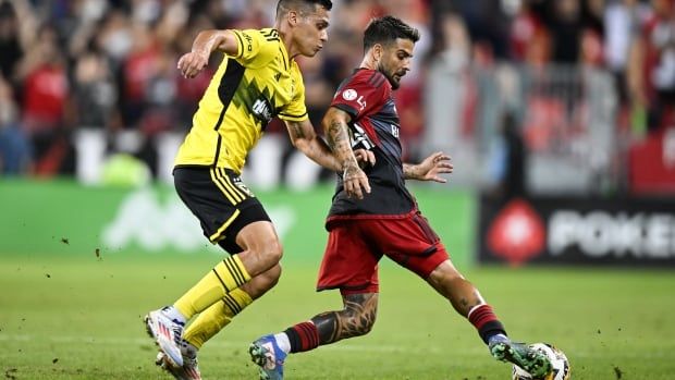 Male soccer player defends ball as an opposing player runs behind him on the soccer pitch. 