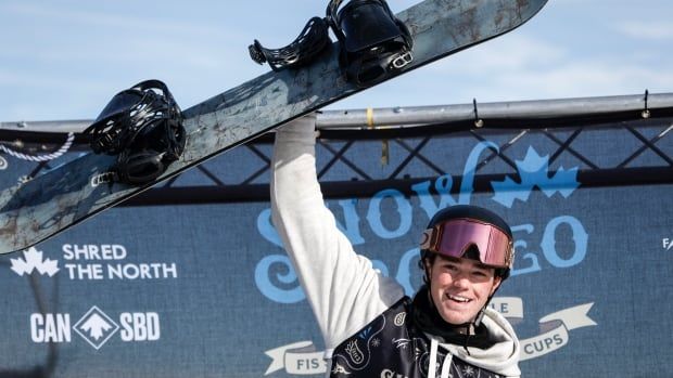 Male snowboard athlete wearing helmet and goggles raises snowboard into the air with one hand in celebration 