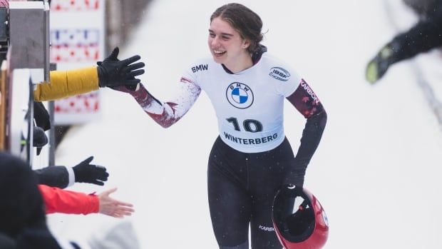 A women's skeleton athlete - wearing a white racing bib and red, white and black skeleton suit - celebrates with fans as she walks on the track while carrying her red helmet.