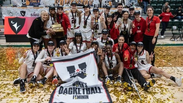 A women's basketball team poses with a banner and a trophy on a basketball court strewn with confetti.