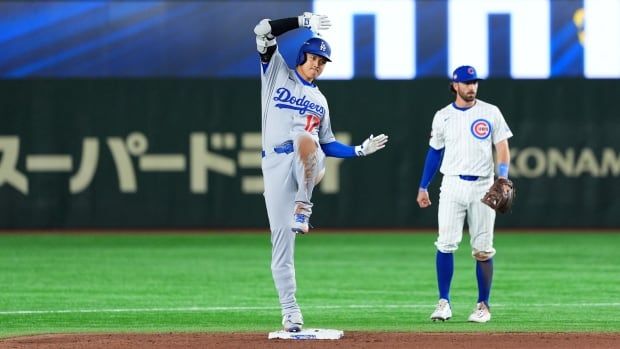 A baseball player stands on second base and celebrates while a defensive player looks on.