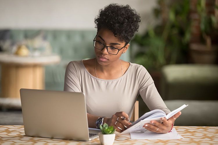 Focused african student looking at laptop holding book doing research