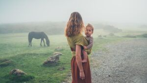 A young mother with her baby in a sling is standing in the fog looking at a horse