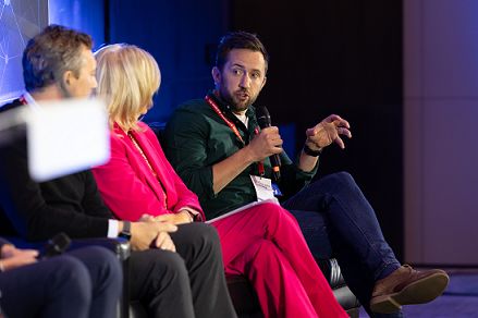 Paul Rowland sitting on stage speaking looking to his right holding a microphone and gesturing with his other hand. Also in shot are a woman in a pink suit (Nina Wright) and a man wearing black (Graham Page) looking at Rowland.