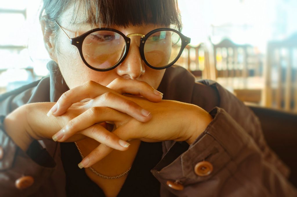 A young woman, wearing glasses and a coat, looks down, her hands folded in front of her mouth