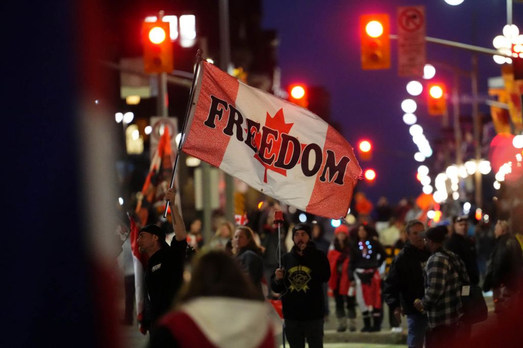 A protester waves a Canadian flag with "freedom" written across it amid a nighttime protest.