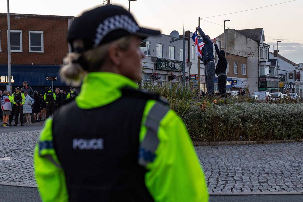 A police offer looks at a town square while in the background, two men in black hoodies raise a Union Jack flag.