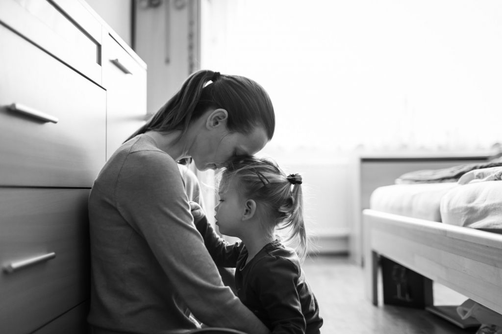 In a black-and-white photo, a mother sits on the floor and bows her head over her young daughter.