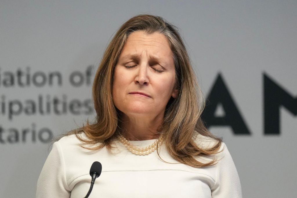 Chrystia Freeland is a white sweater and pearls stands in front of a microphone with her eyes shut