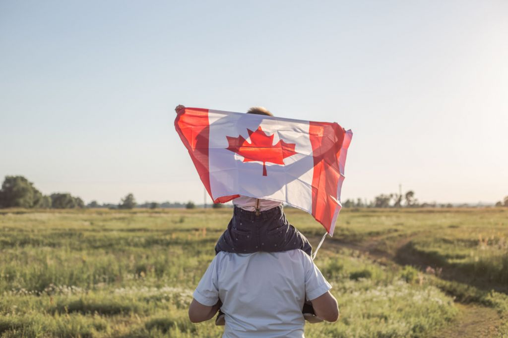 Pictured from behind, a man carries a young child draped in the Canadian flag on his shoulders