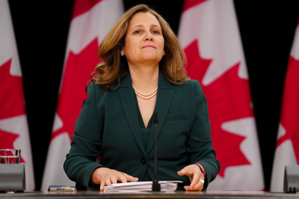 A photo of Chrystia Freeland, in front of a row of Canadian flags, wearing a green suit and holding a stack of papers