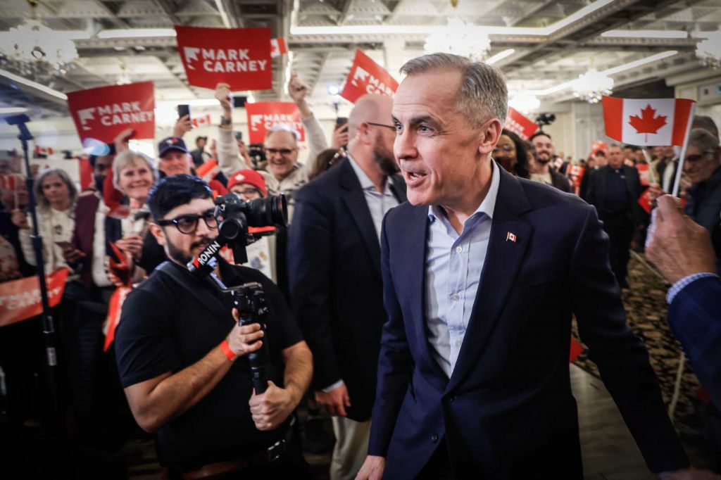 A photo of Mark Carney looking surprised while people around him hold up small Canadian flags and red signs with his name