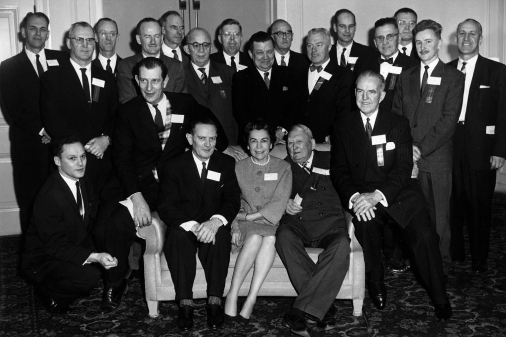 A black-and-white photo of a group of men in suits, several rows deep, crowding around a sole woman sitting on a couch with two other men.