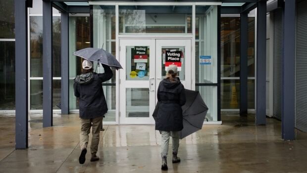 People in rain jackets carrying umbrellas walk into a building marked with signs that say "voting place".