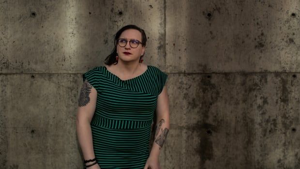 A woman in a green dress stands against a concrete wall. 