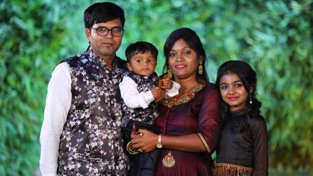 An Indian famiy of four is pictured against a green leafy backdrop. A man stands on the left hand side, a woman holds a toddler boy, and an older girl stands on her right.