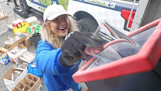 A woman pushes a glass into the mouth of a glass-crushing machine. There is a pile of household glass on the ground behind her.