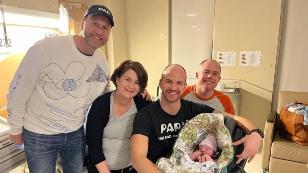 Four adults pose for a photo in a hospital room with a newborn baby in a car seat