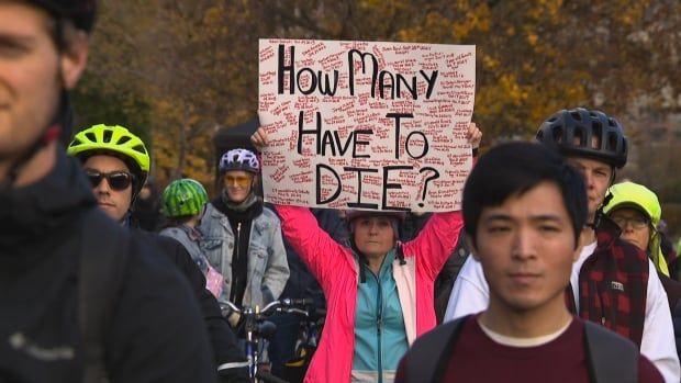 A woman holds up a sign at a rally that says 'How many have to die' while surrounded by people wearing bike helmets. 