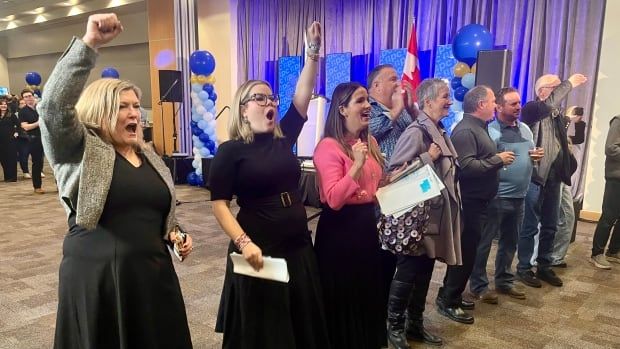 A line of people dressed in smart clothing cheer and pump their fists in the air in a banquet room.