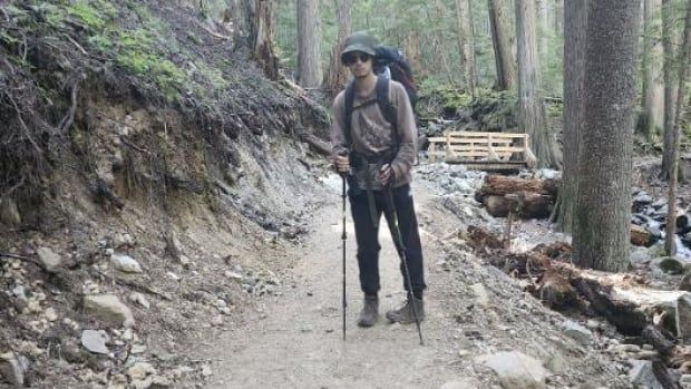 A man in a hat, with a grey shirt, hiking boots, poles and a hiking backpack. 